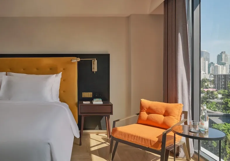 Edge of the City with a mustard tufted headboard, a brass reading light and an orange armchair against the window wall