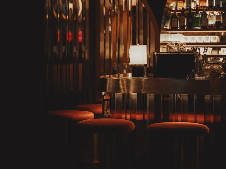The counter at OPEN BAR after dark, orange stools along a fluted bar front with the back-bar shelves lit behind