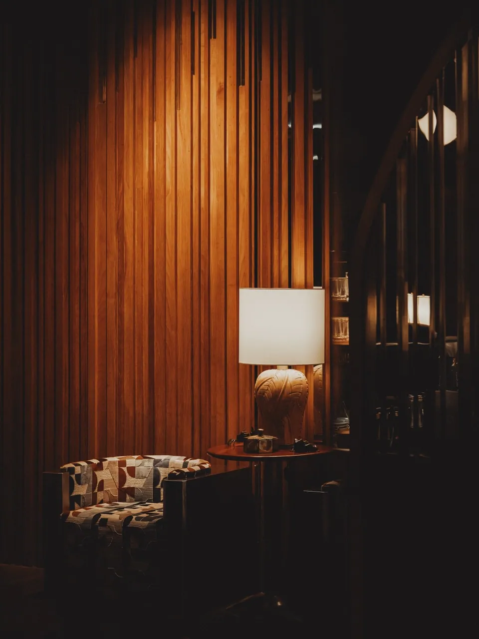 A lamp-lit table and patterned armchair against the fluted timber wall in the lounge at OPEN BAR