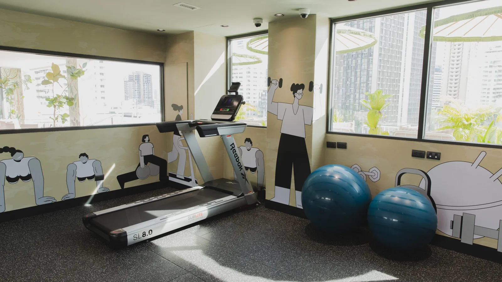 The gym at Public House Bangkok: a treadmill and two exercise balls on a rubber floor, an illustrated wall of figures lifting weights behind them and the Sukhumvit skyline through the windows