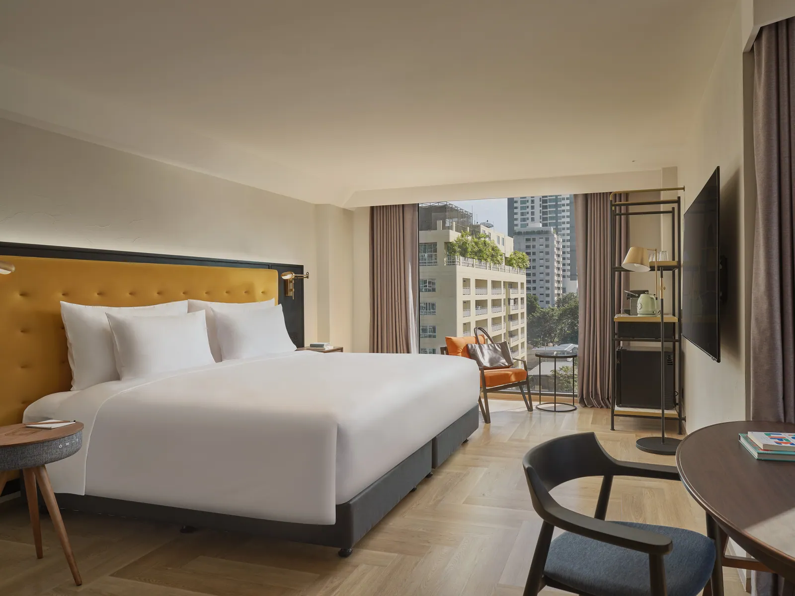 A corner guest room at Public House Bangkok with a yellow upholstered headboard and a window onto the Sukhumvit skyline