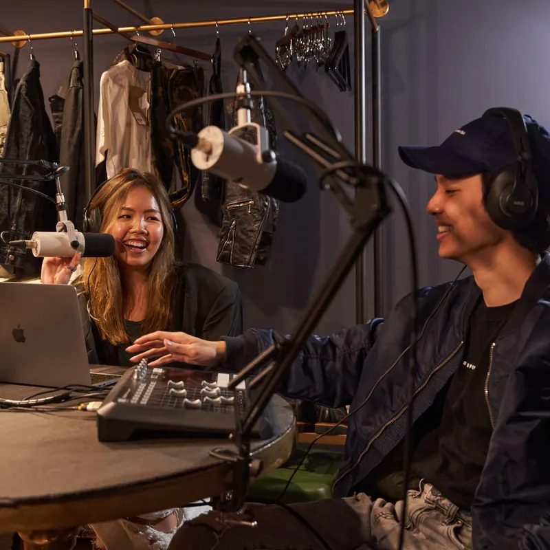 Two hosts recording in The Podcast Lounge at Public House Bangkok, in headphones at the microphones with a laptop and mixer on the stone table