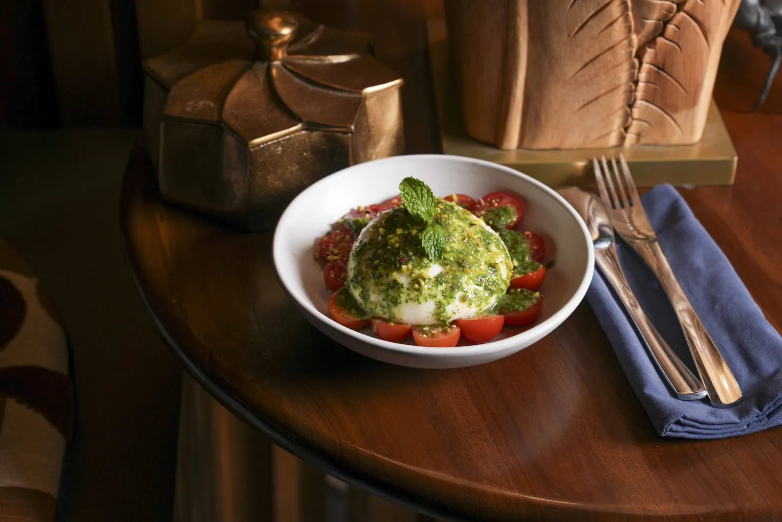 Burrata with tomatoes and herb dressing in a bowl at SWD, on a round wooden table beside a leather banquette