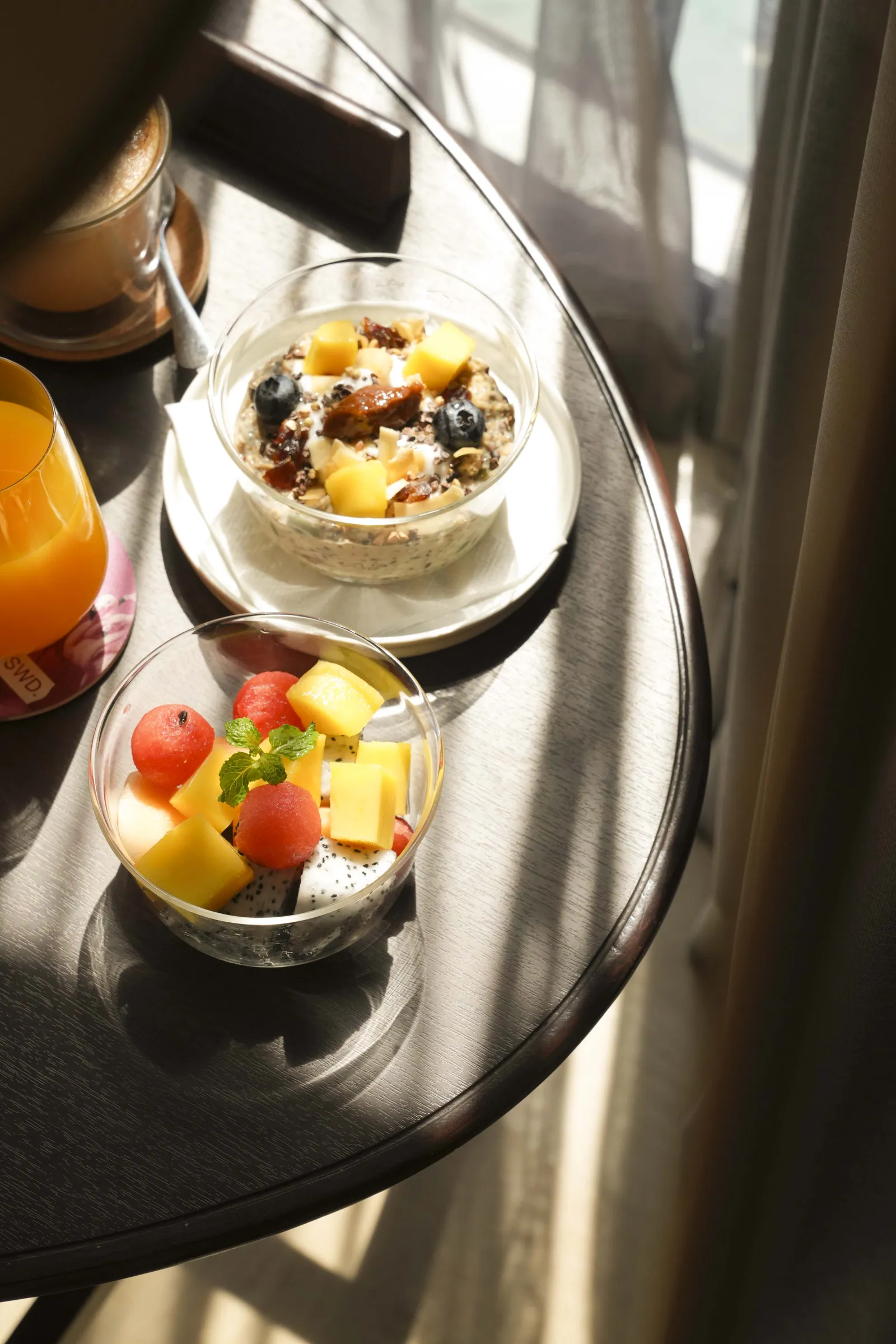 A round table by a curtained window at SWD in the morning sun: a glass of orange juice, a coffee, a bowl of granola with mango and blueberries, and a cup of cut watermelon, mango and dragon fruit with mint