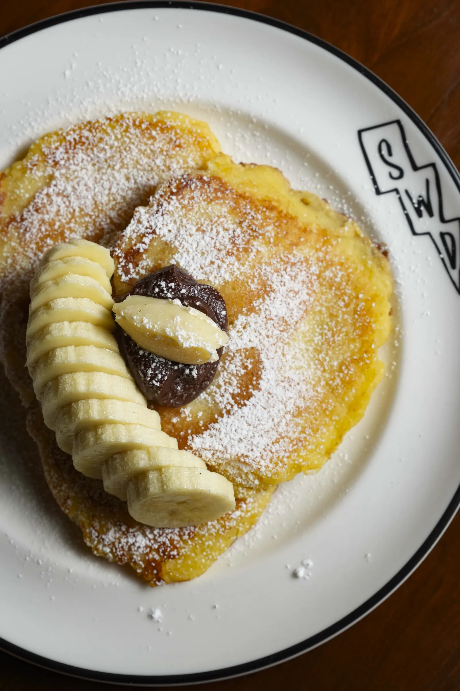 A pancake at SWD with sliced banana, quenelles of custard and icing sugar, on a branded SWD plate
