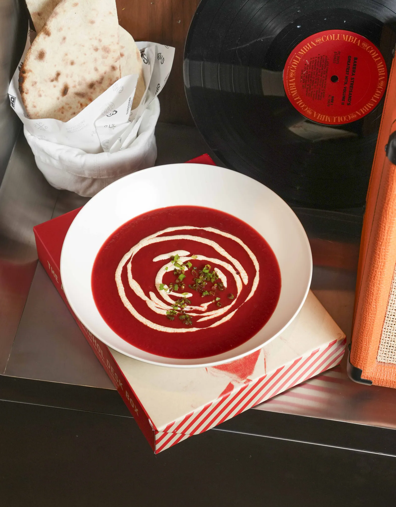 A bowl of deep red soup swirled with cream at SWD, with flatbread in paper and a vinyl record beside it